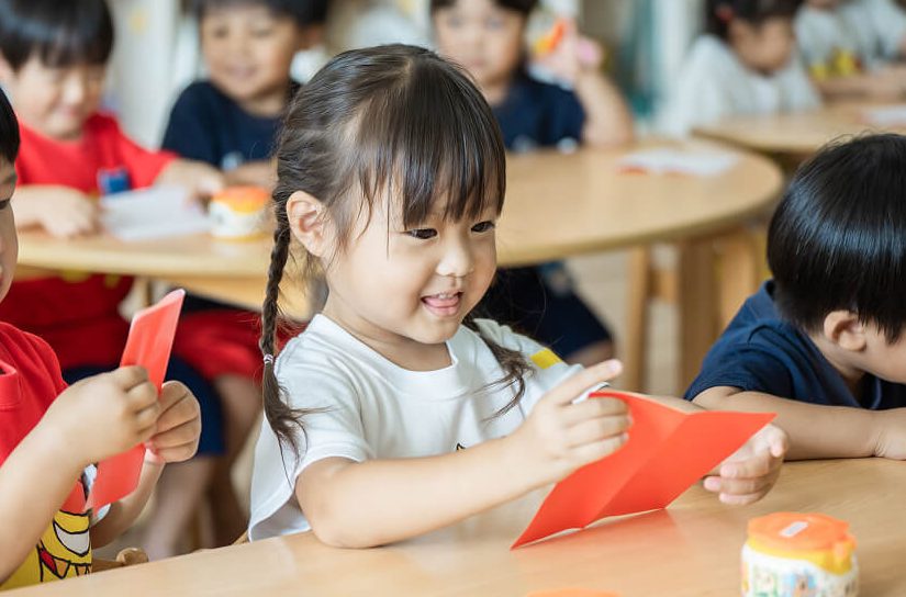 折り紙をする園児: 教室の机で、先生や友達と楽しそうに赤い折り紙を折る女の子。