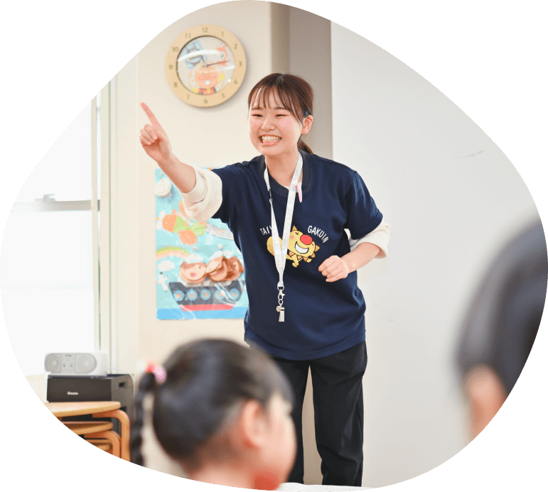 学院のTシャツを着て笑顔で上を指差す先生
